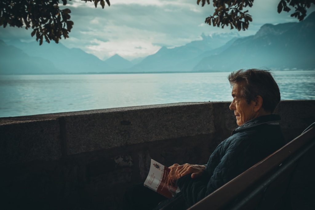 Senior man sitting on bench holding book looking out at water
