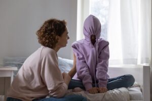 Mom sits on bed and tries to talk to child after learning signs her teen is using drugs, but her teen is hiding in her hoodie